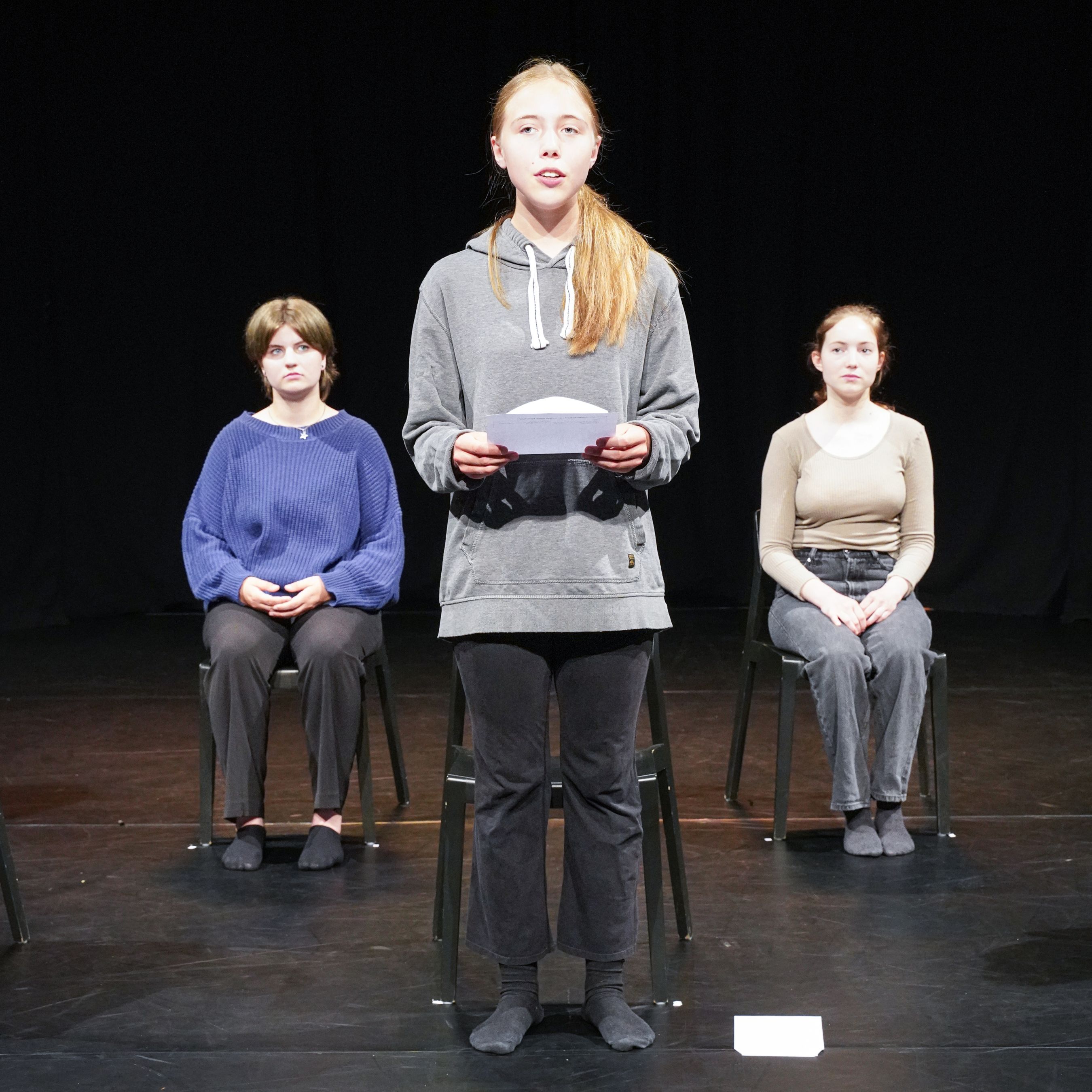 Eine junge Frau steht vorne auf der Bühne und liest einen Brief. Zwei Frauen sitzen hinter ihr und schauen zu ihr.
